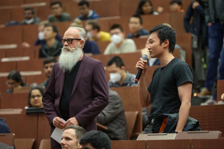 Moderator and a participant stand in the lecture hall and direct their questions to the presenters.